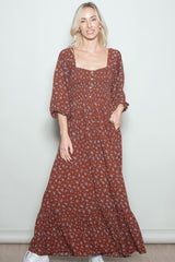 woman wearing a rust floral maxi dress with a square neckline and puff sleeves, standing with one hand in the pocket
