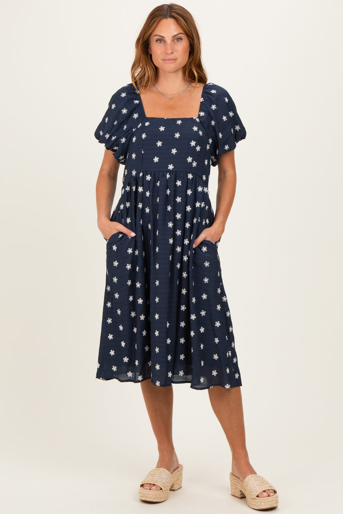 woman wearing a navy blue midi dress with white floral patterns and puff sleeves, standing with hands in pockets