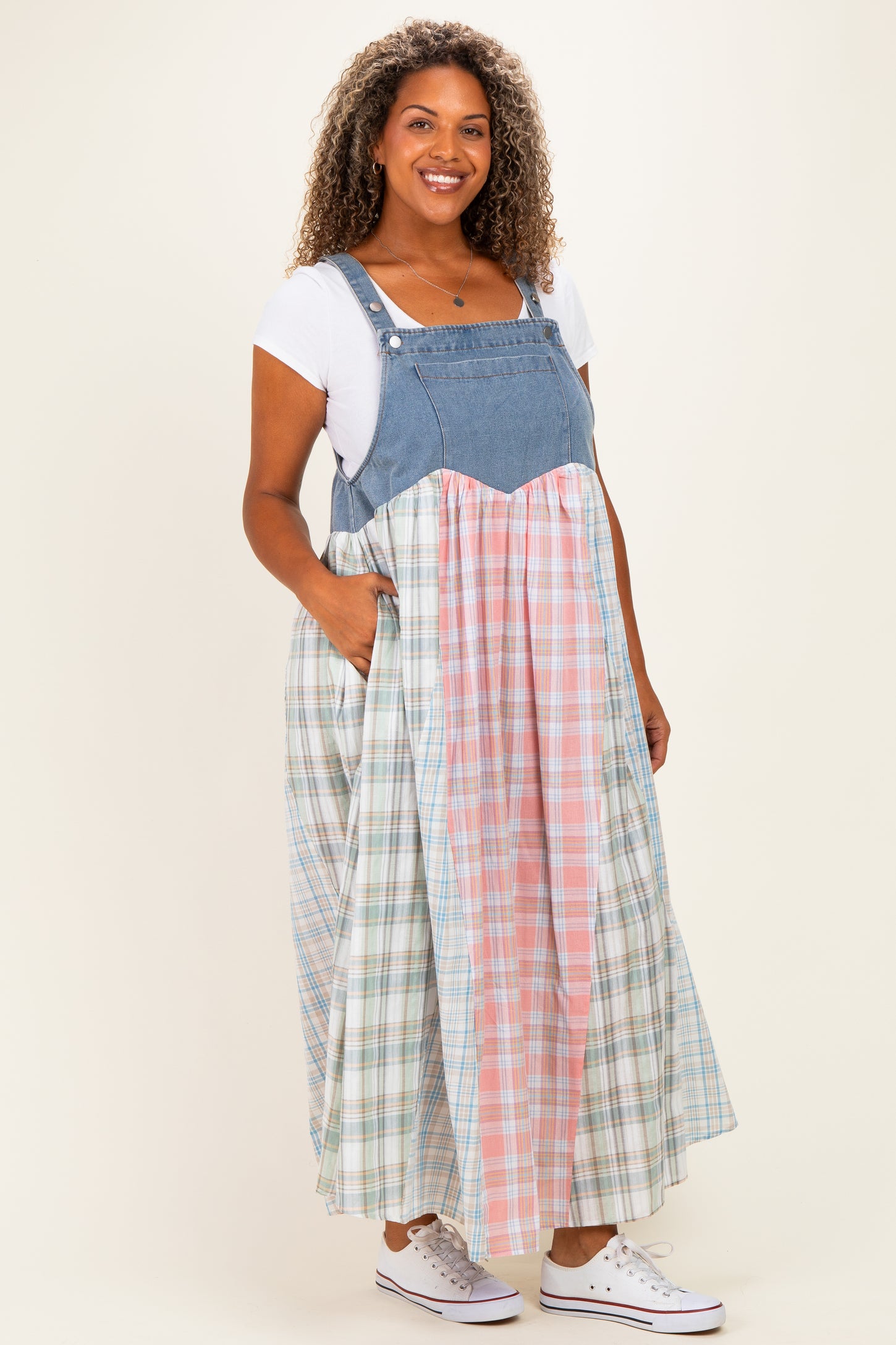 woman wearing a plus size plaid maxi dress with denim straps and pocket details, styled with white sneakers