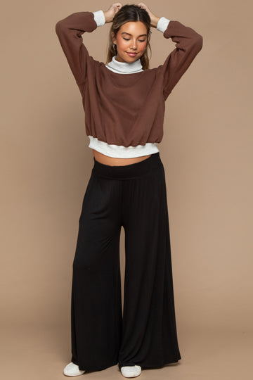 woman wearing a brown cropped sweatshirt over a gray turtleneck and black maternity wide-leg pants with a visible baby bump