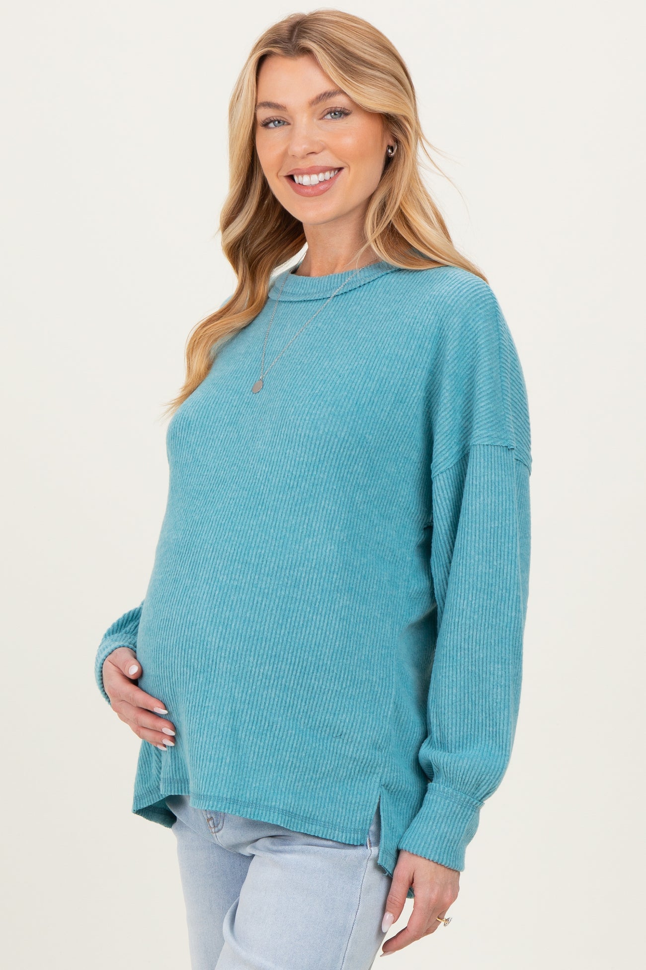 woman wearing a teal ribbed maternity sweater and holding her baby bump, paired with light wash jeans
