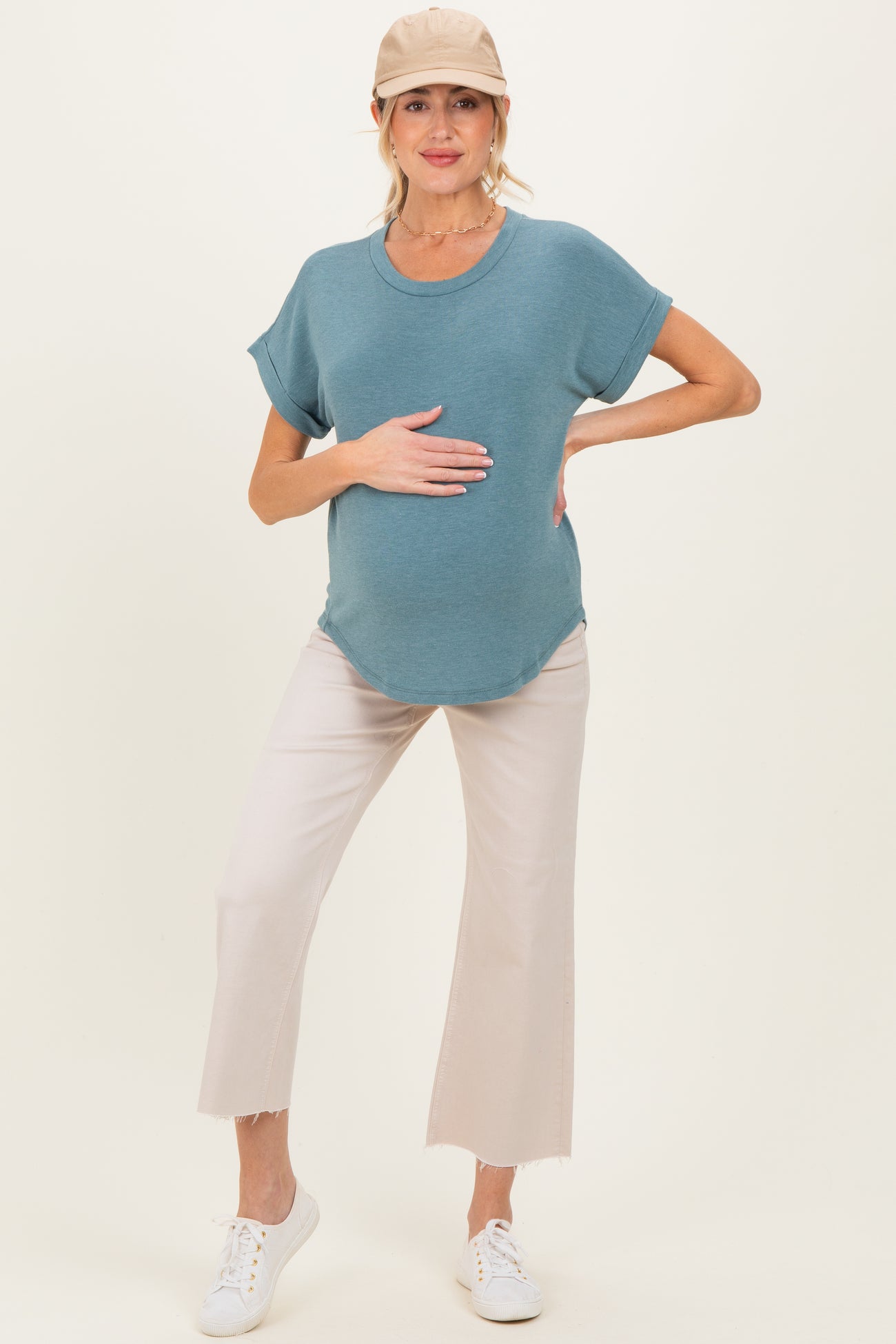 woman wearing a teal maternity top and light beige cropped pants, holding her baby bump