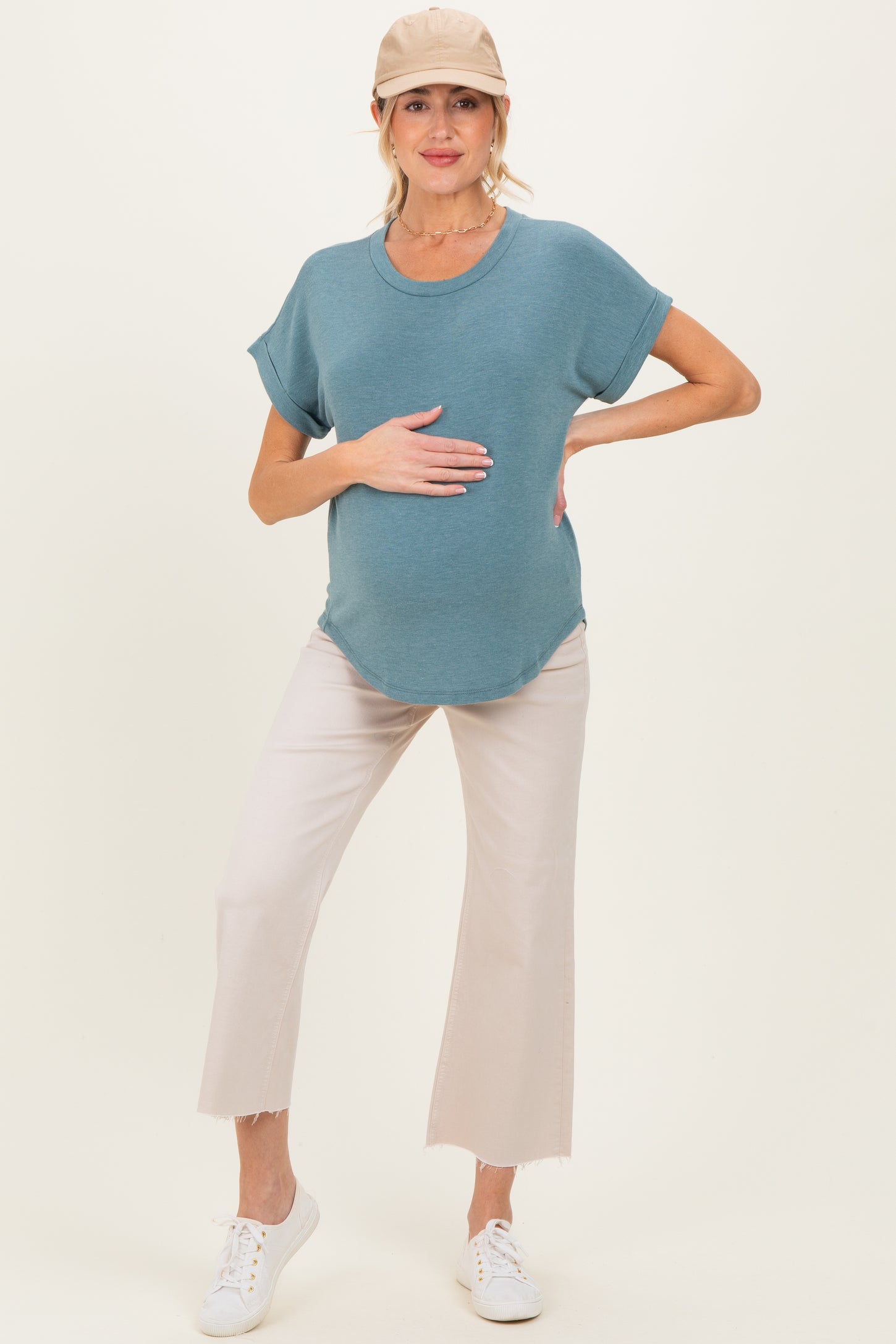 woman wearing a teal maternity top and light beige cropped pants, holding her baby bump