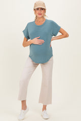 woman wearing a teal maternity top and light beige cropped pants, holding her baby bump