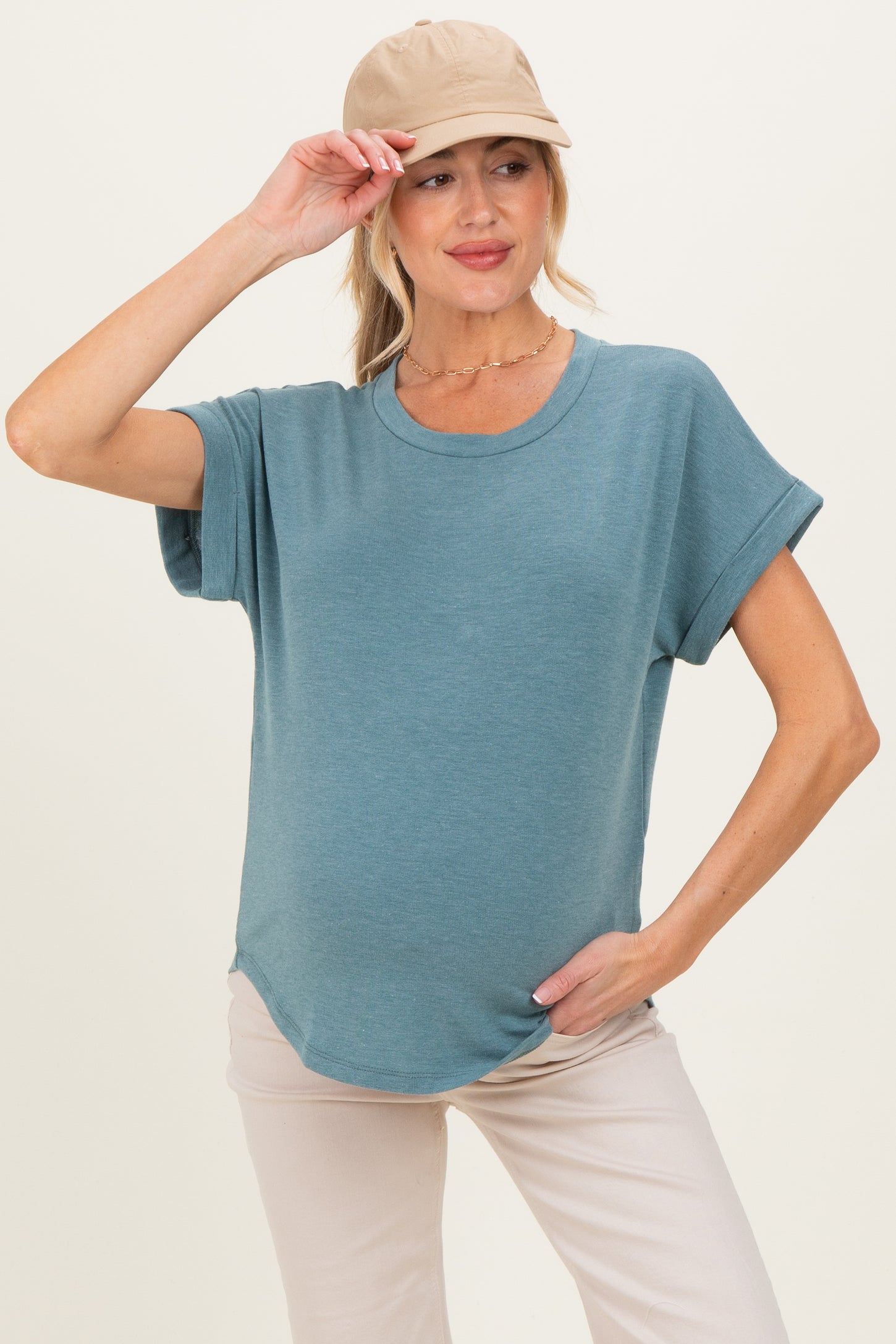 woman wearing a blue short sleeve maternity top with a relaxed fit and holding a beige cap, showing her baby bump