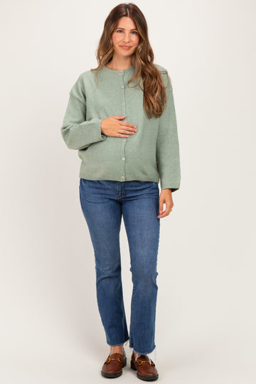 woman wearing a green maternity cardigan and blue jeans, gently cradling her baby bump