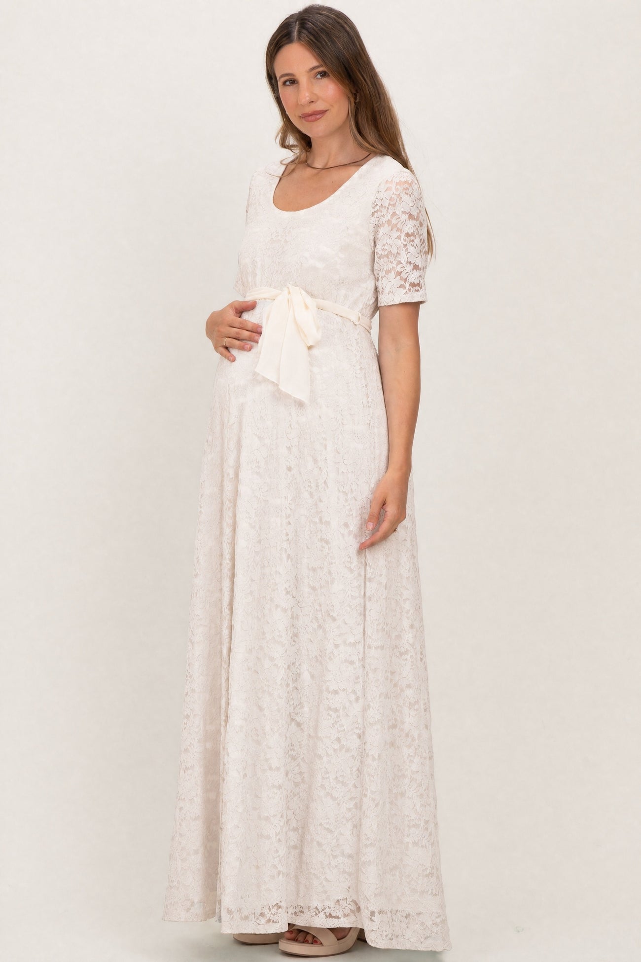 woman wearing a cream lace maternity gown with short sleeves and a ribbon belt, gently holding her baby bump