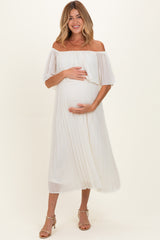 woman wearing a white off-the-shoulder maternity dress, gently holding her baby bump with pleated detail