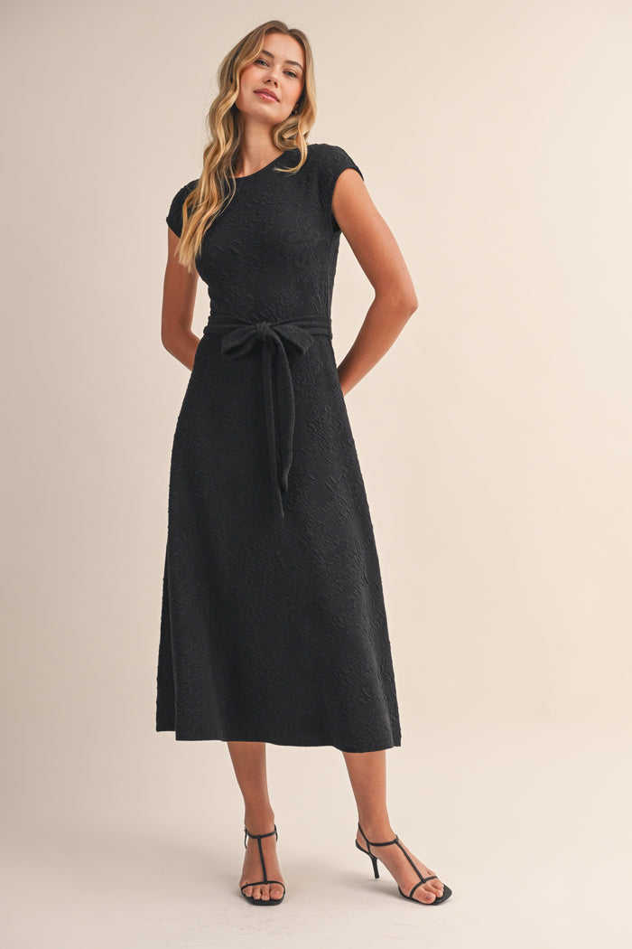 woman wearing a black mid-length dress with a fitted bodice and a tie belt, styled with strappy sandals