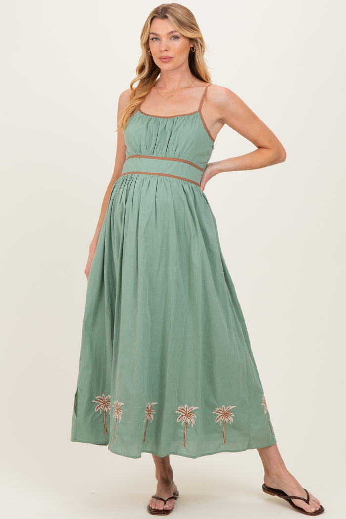 woman wearing a green maternity maxi dress with a cinched waist and palm tree embroidery at the hem