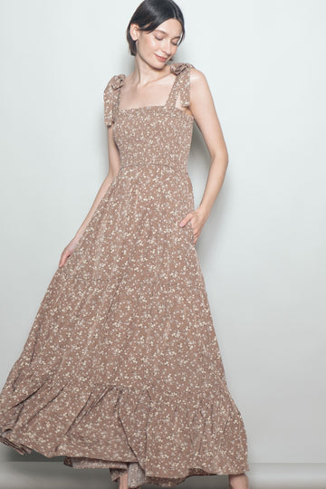 woman wearing a brown floral maxi dress with square neckline and bow tie straps, smiling and posing with one hand in pocket