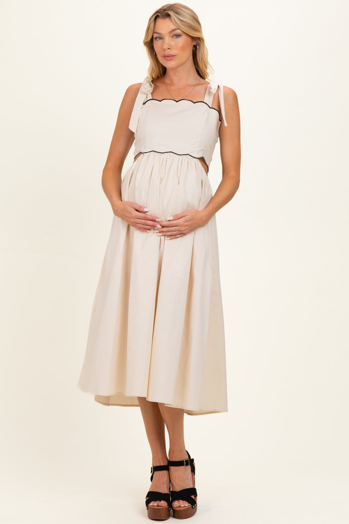 woman wearing a cream maternity dress with scalloped straps, holding her baby bump and standing in a light studio