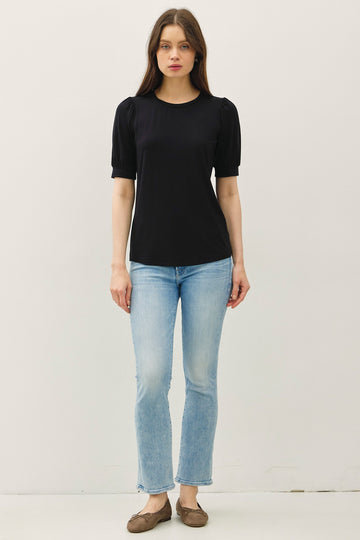 woman wearing a black short sleeve top with puff sleeves and light blue flared jeans, standing in a neutral setting
