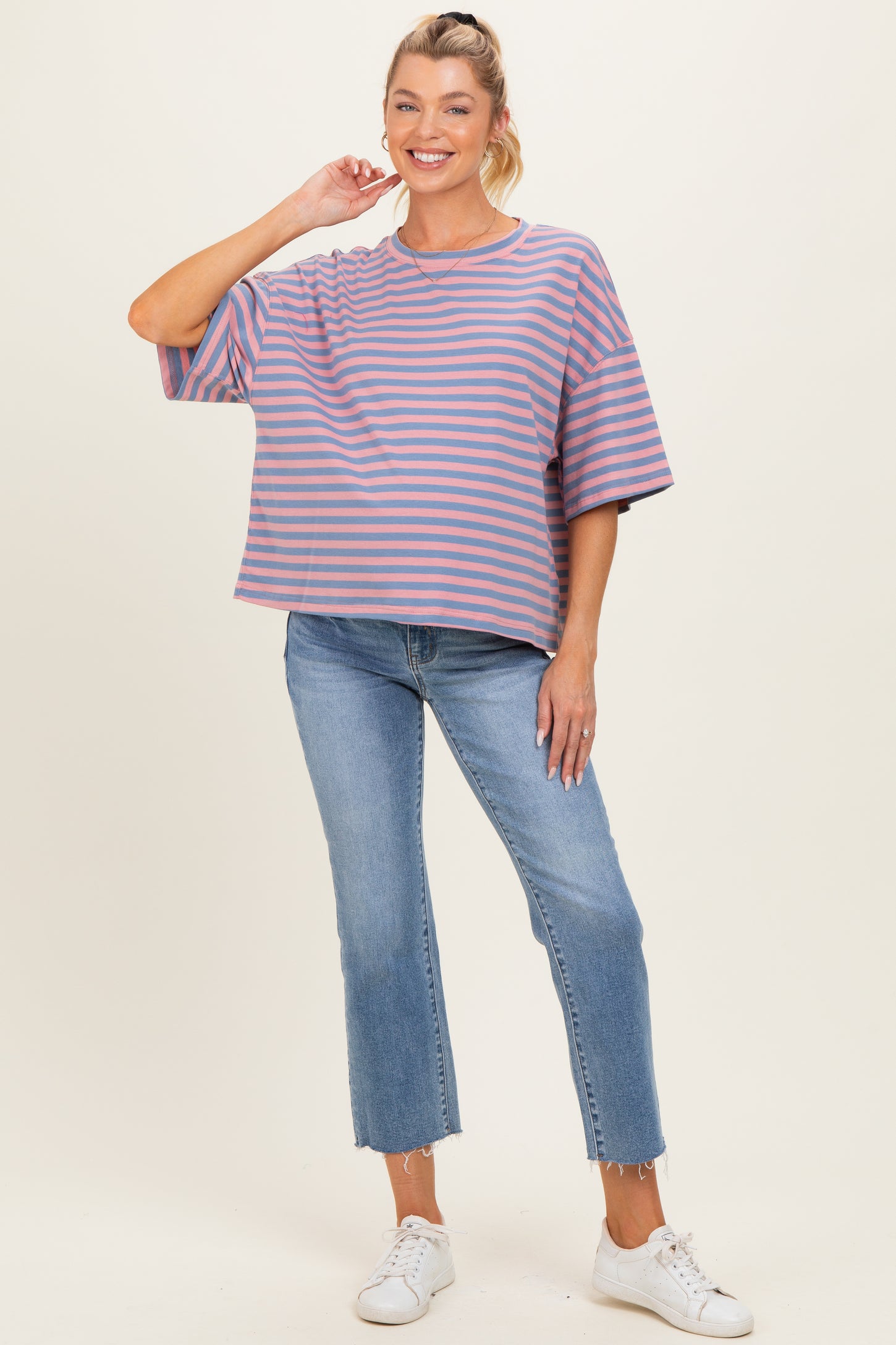 woman wearing a striped purple and pink maternity top with loose fit and blue jeans, smiling and standing with one hand on her hip