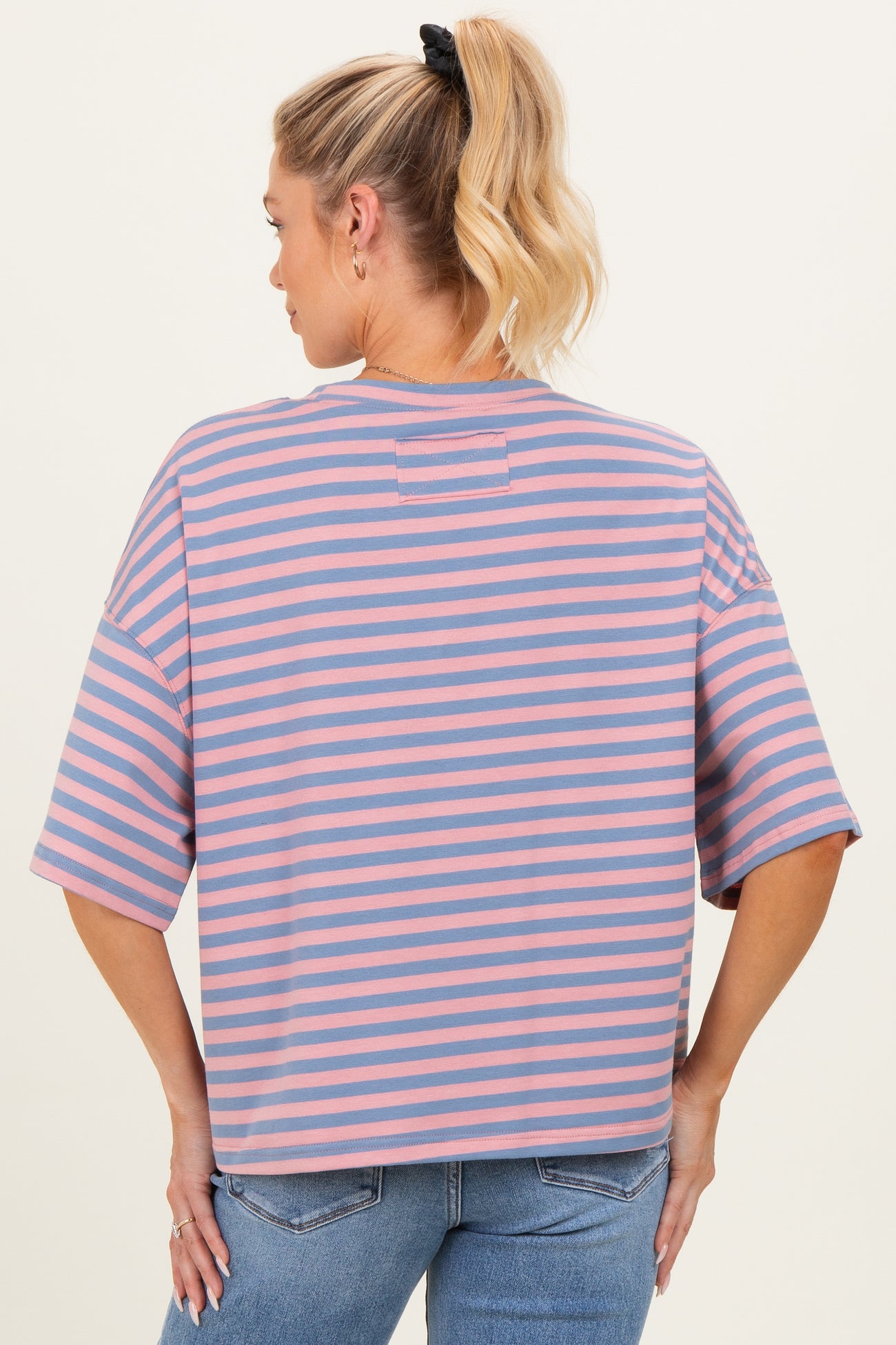woman wearing a striped maternity top in light pink and blue, viewed from the back, with loose short sleeves and relaxed fit