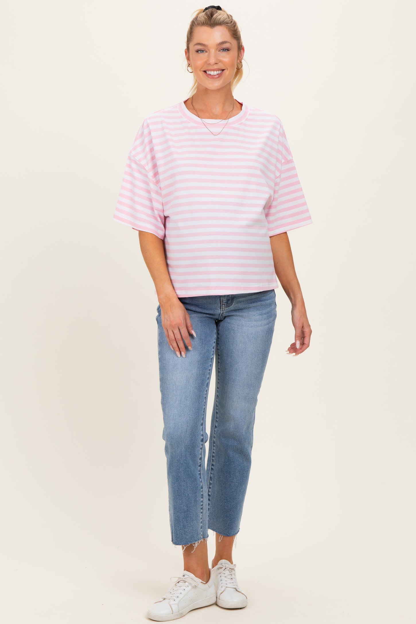 woman wearing a pink and white striped maternity top paired with light wash jeans and white sneakers