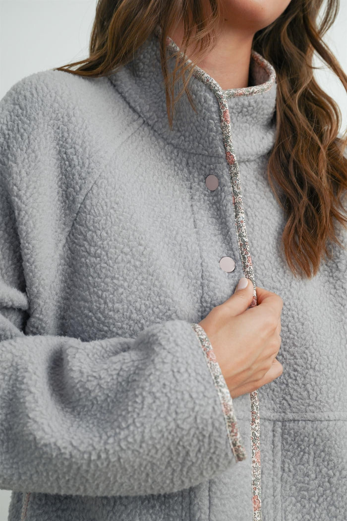 woman wearing a gray fleece jacket with a high collar and decorative trim, holding it closed with one hand