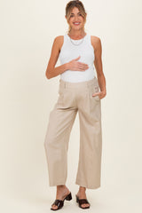 woman wearing a white tank top and beige wide-leg maternity pants, gently holding her baby bump