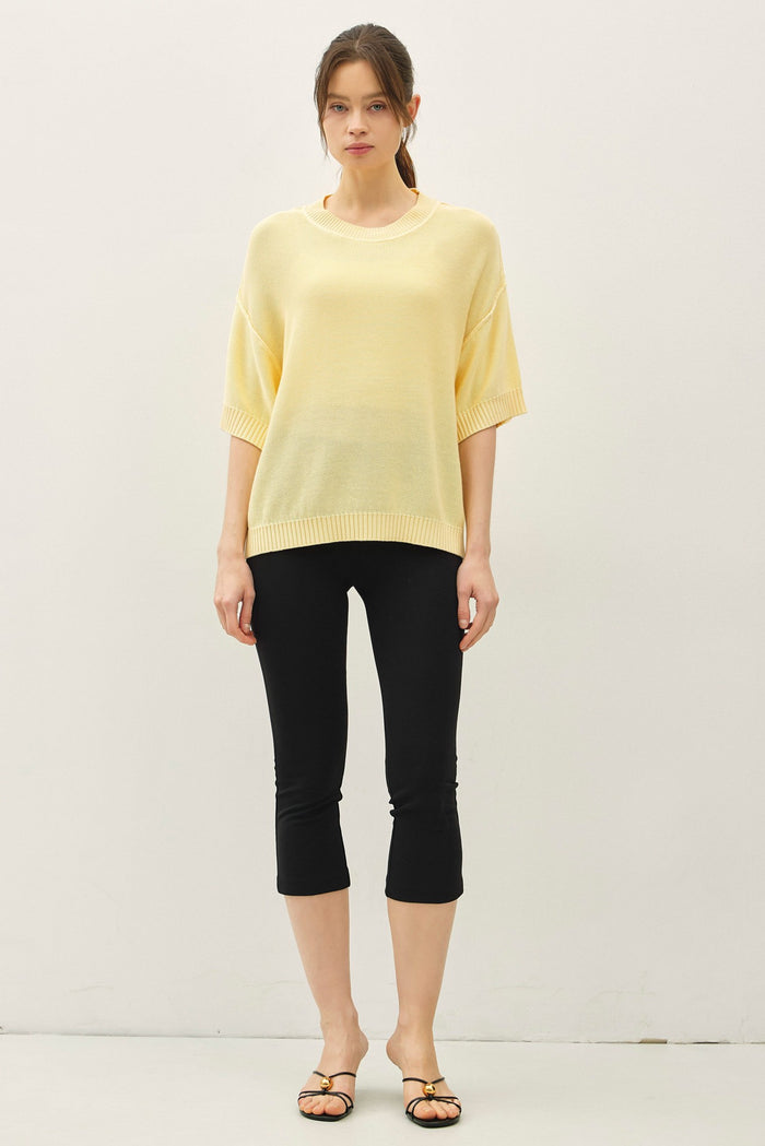 woman wearing a light yellow oversized sweater and black capri leggings, standing against a neutral background