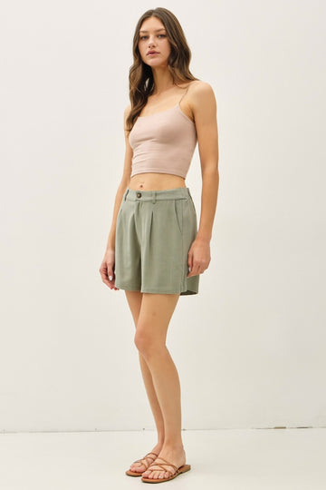 woman wearing a beige crop top and green high-waisted shorts, standing with a neutral background