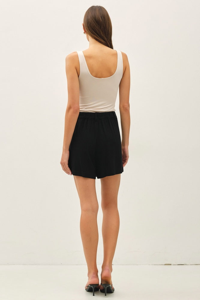 woman wearing a beige tank top and black shorts, viewed from the back, standing against a plain background