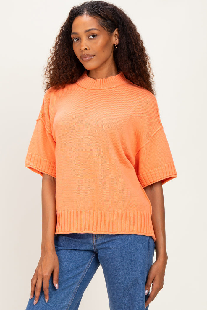 woman wearing an oversized bright orange short sleeve sweater with a ribbed hem and standing in front of a neutral background