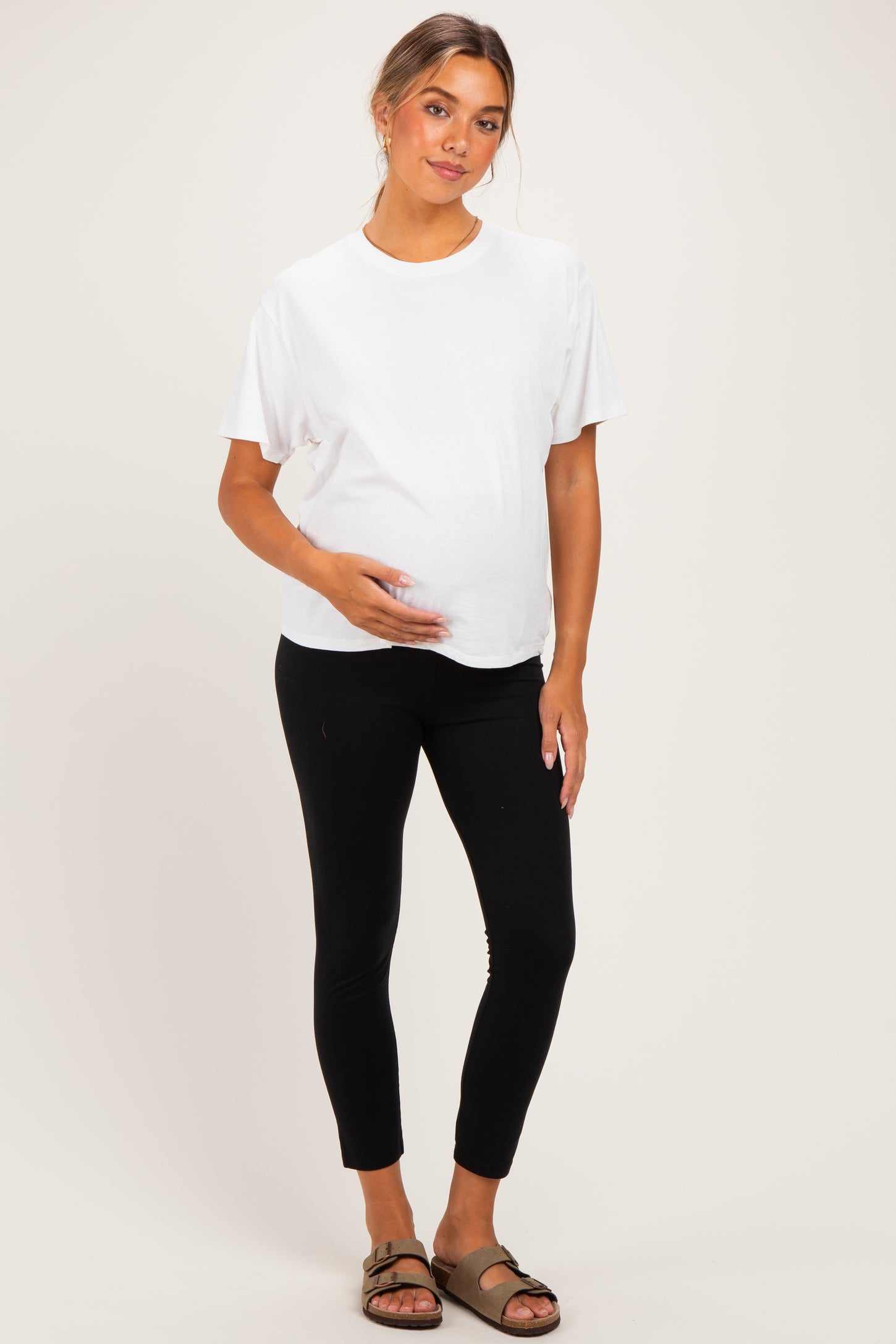 woman wearing a white short sleeve maternity top and black leggings, gently holding her baby bump