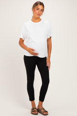 woman wearing a white short sleeve maternity top and black leggings, gently holding her baby bump