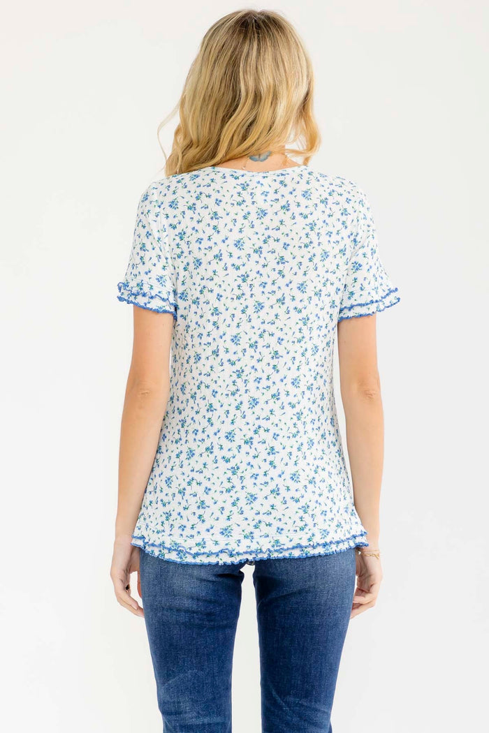 woman wearing a white short sleeve top with blue floral print and ruffle details, view from the back