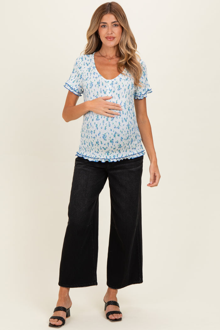 woman wearing a light blue floral maternity top with ruffle details and black wide-leg pants, holding her baby bump
