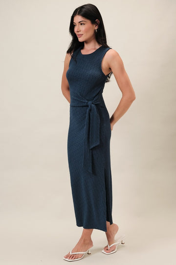 woman wearing a navy blue sleeveless midi dress with a tie waist and side slit, standing against a neutral background