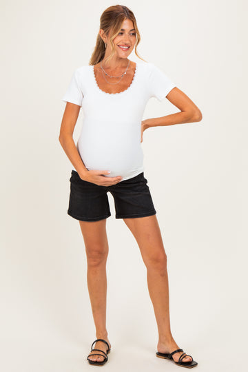 woman wearing a white short sleeve maternity top and black denim shorts, cradling her baby bump