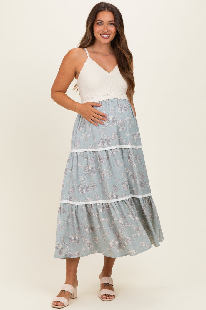 woman wearing a maternity maxi dress with a white knitted bodice and a blue floral tiered skirt, holding her baby bump