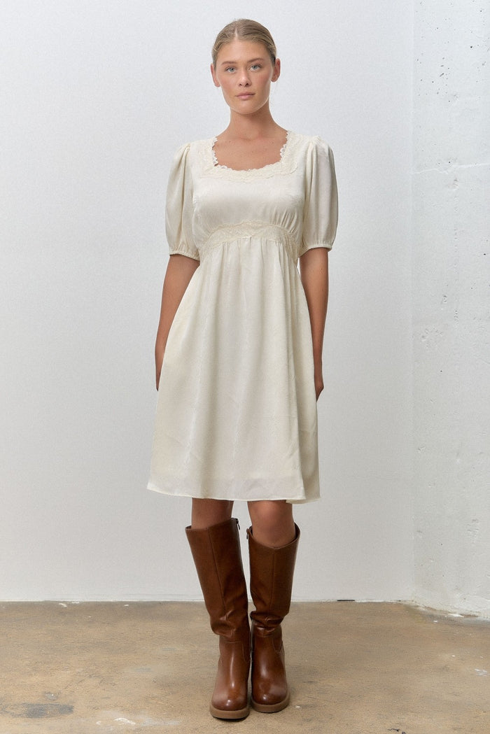 woman wearing a cream short puff sleeve dress with a square neckline and holding brown knee-high boots