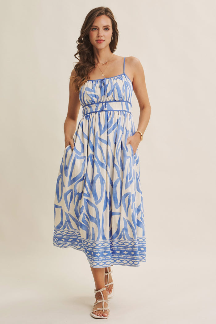 woman wearing a blue and white patterned midi dress with spaghetti straps and pockets, styled with strappy sandals