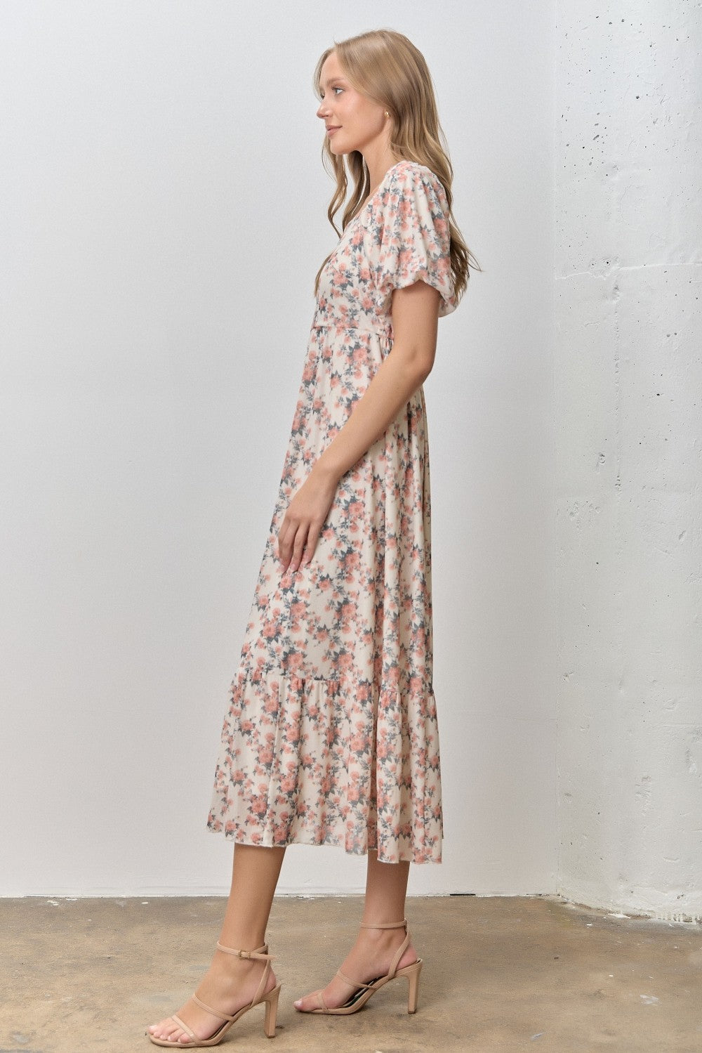 woman wearing a light pink floral midi dress with short sleeves and a ruffled hem, standing against a white wall