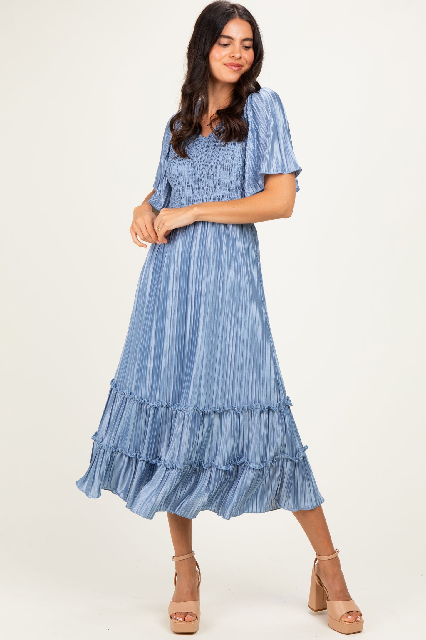 woman wearing a blue pleated midi dress with ruffle details and short sleeves, standing with a smile and wearing heeled sandals