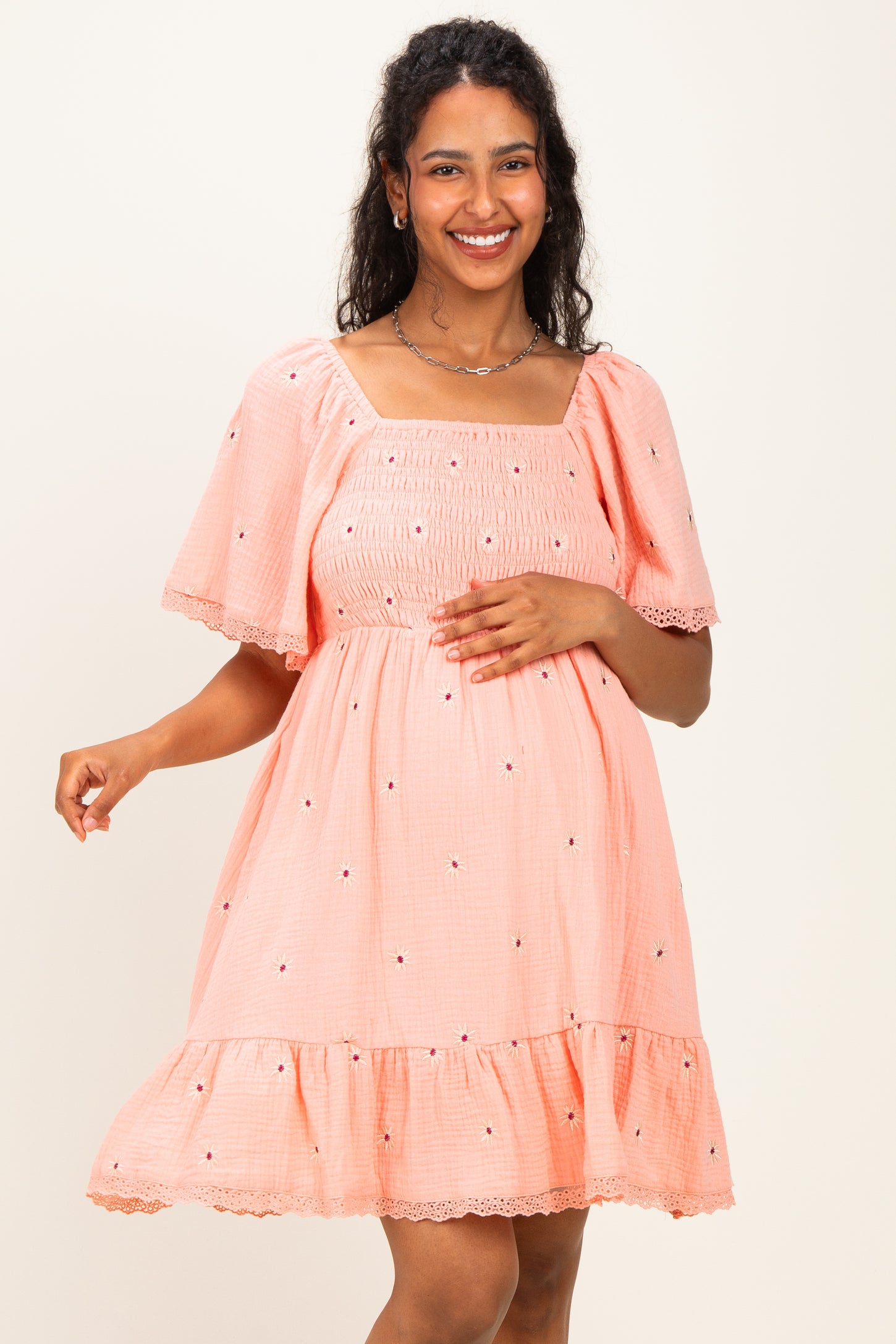 woman wearing a pink embroidered maternity dress with short sleeves and a square neckline, holding her baby bump