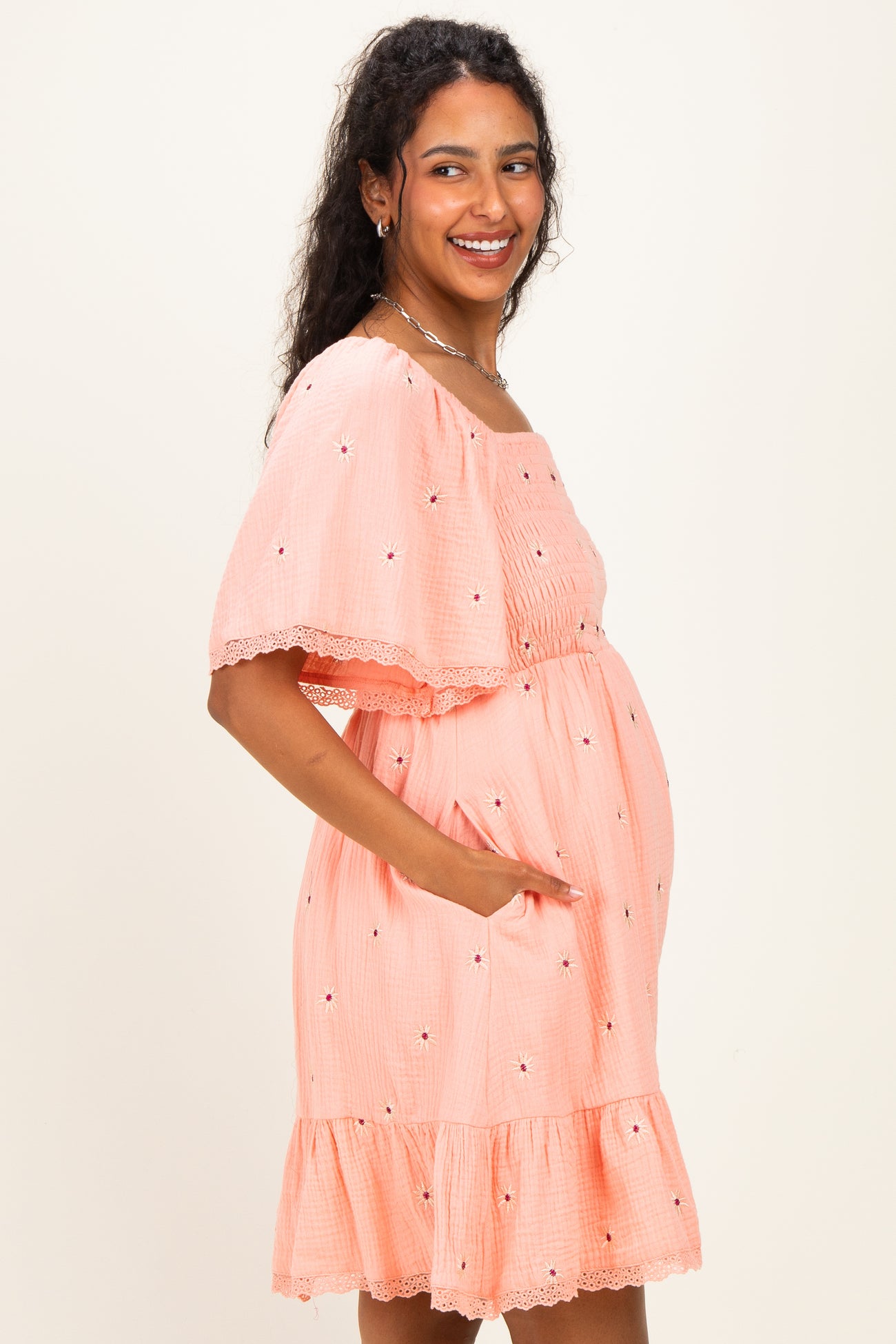 woman wearing a coral maternity dress with a ruffled hem, short sleeves, and floral embroidery, showcasing her baby bump