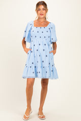 woman wearing a light blue maternity dress with floral embroidery and short flutter sleeves, featuring pockets