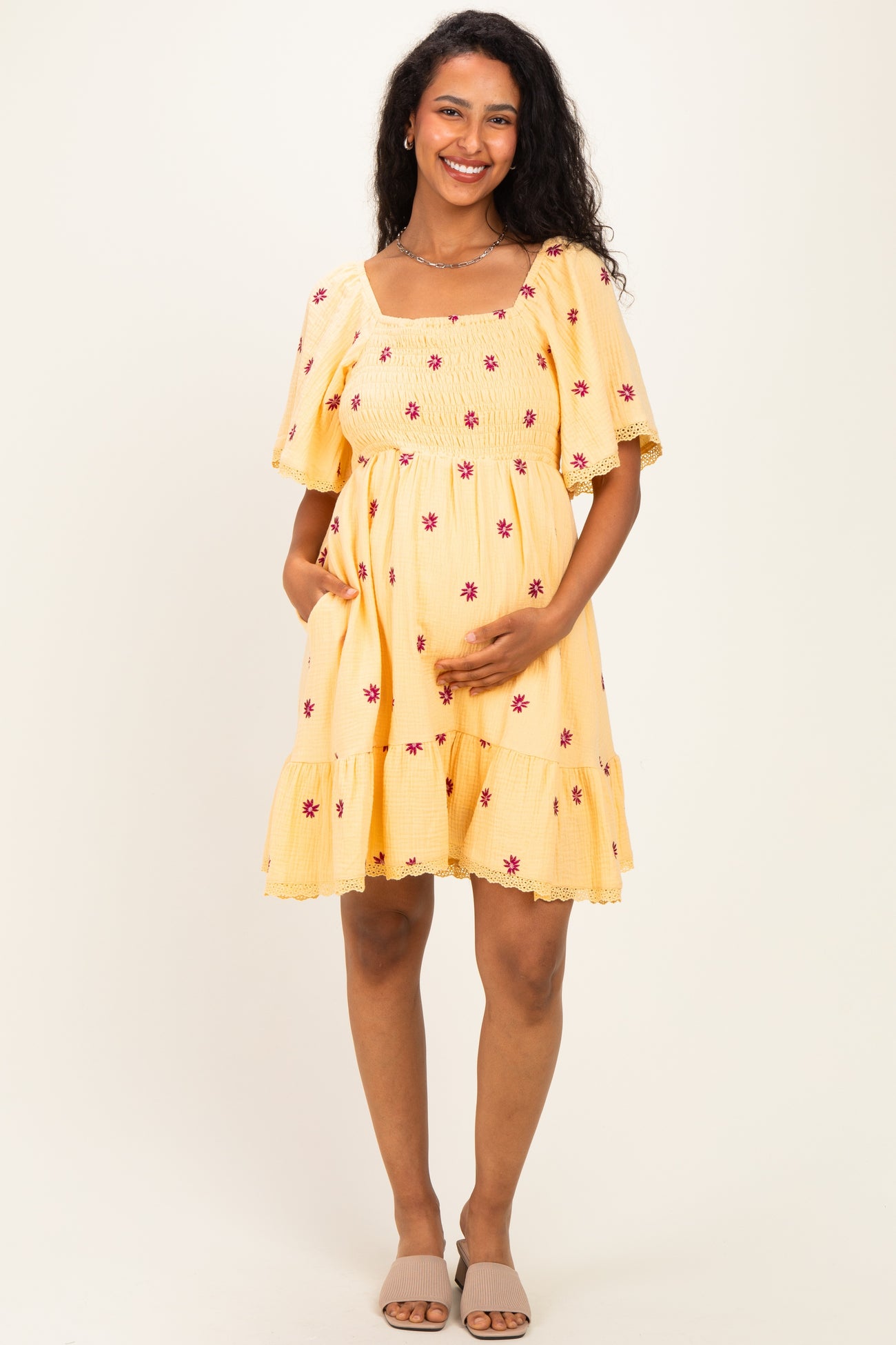 woman wearing a yellow maternity dress with floral embroidery and a tiered hem, holding her baby bump