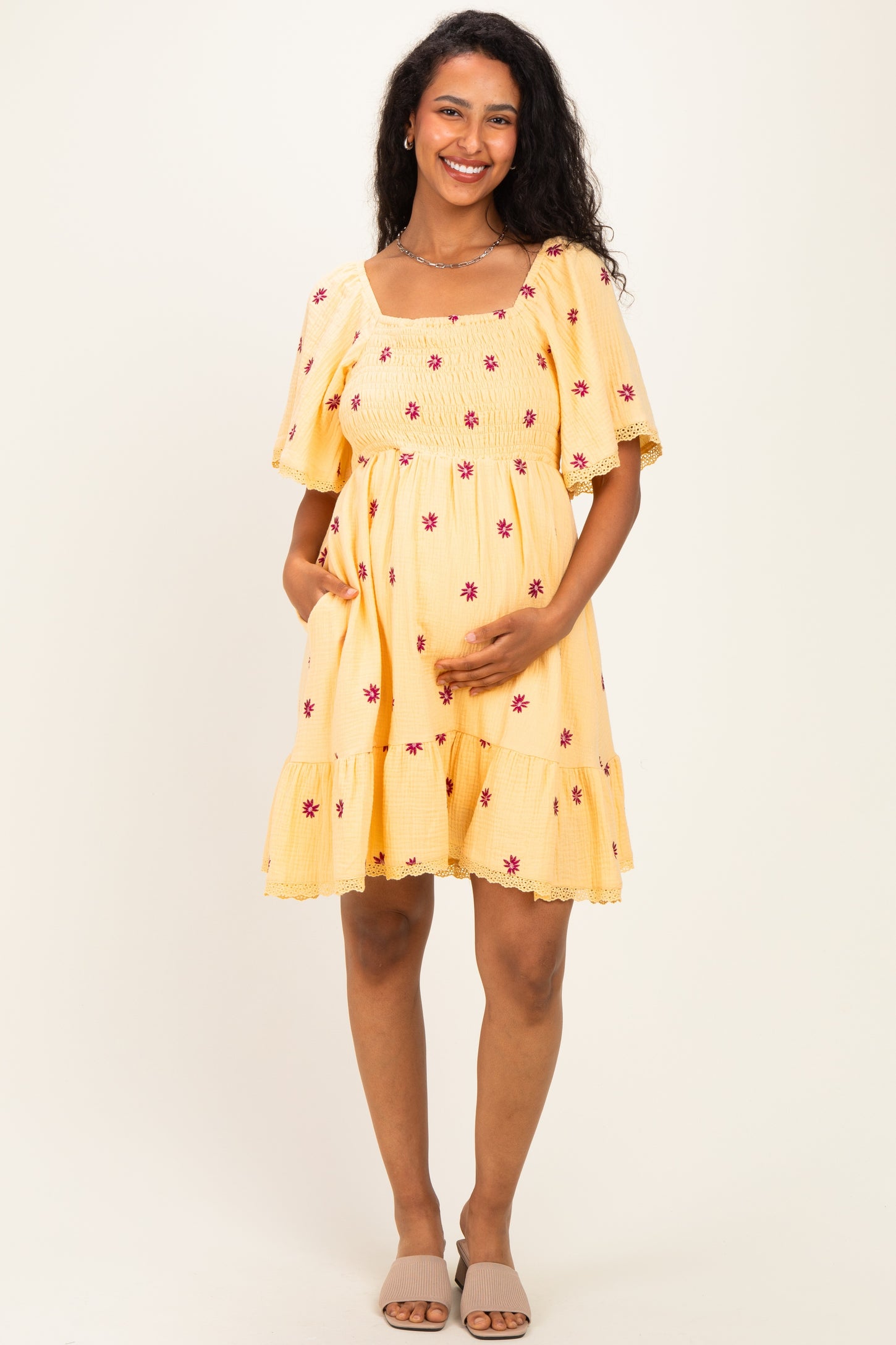 woman wearing a yellow maternity dress with floral embroidery and a tiered hem, holding her baby bump