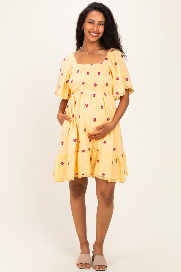woman wearing a yellow maternity dress with floral embroidery and a tiered hem, holding her baby bump