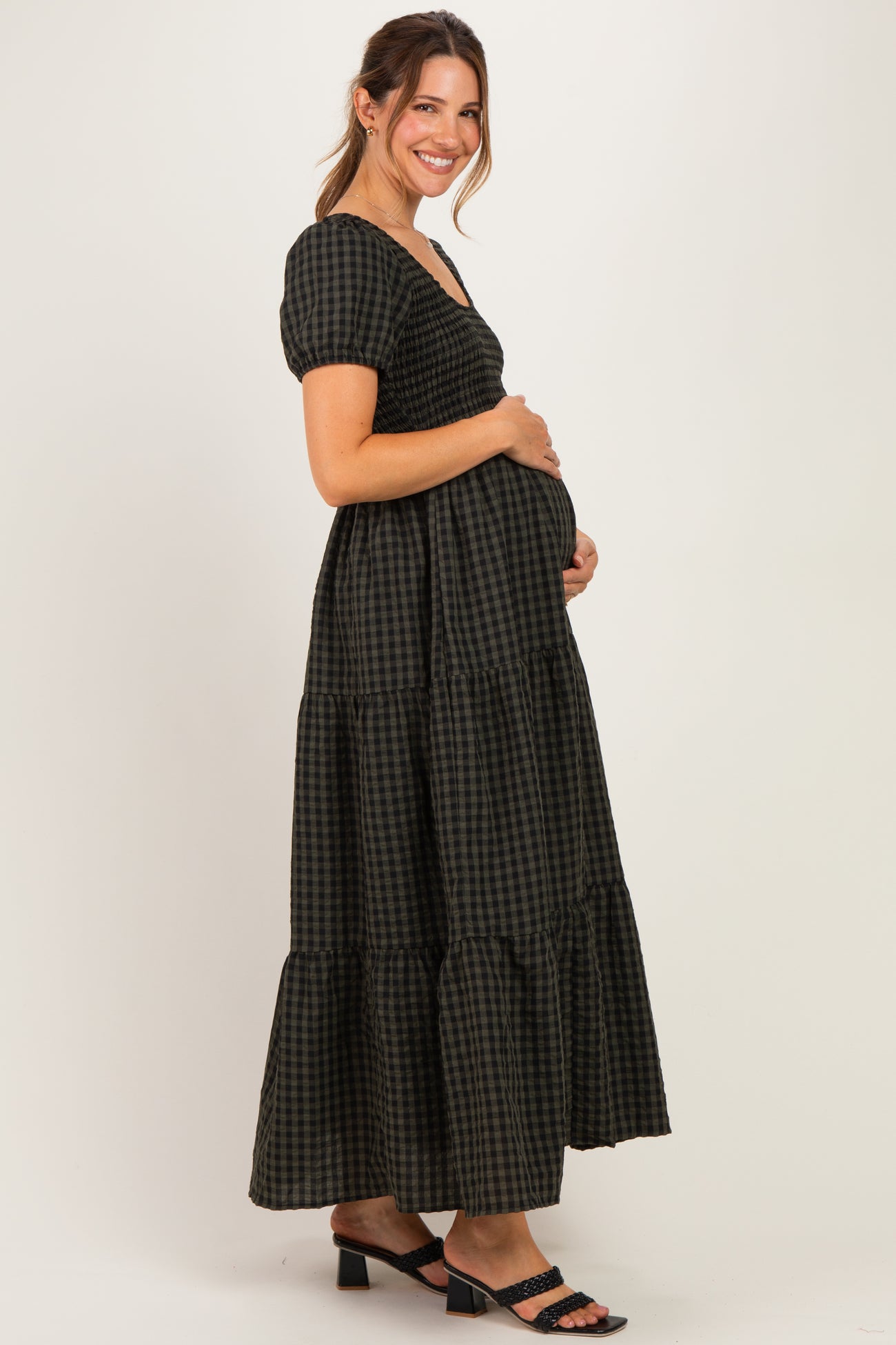 woman wearing a black and green gingham check maternity dress with short puff sleeves and a tiered skirt, cradling her baby bump