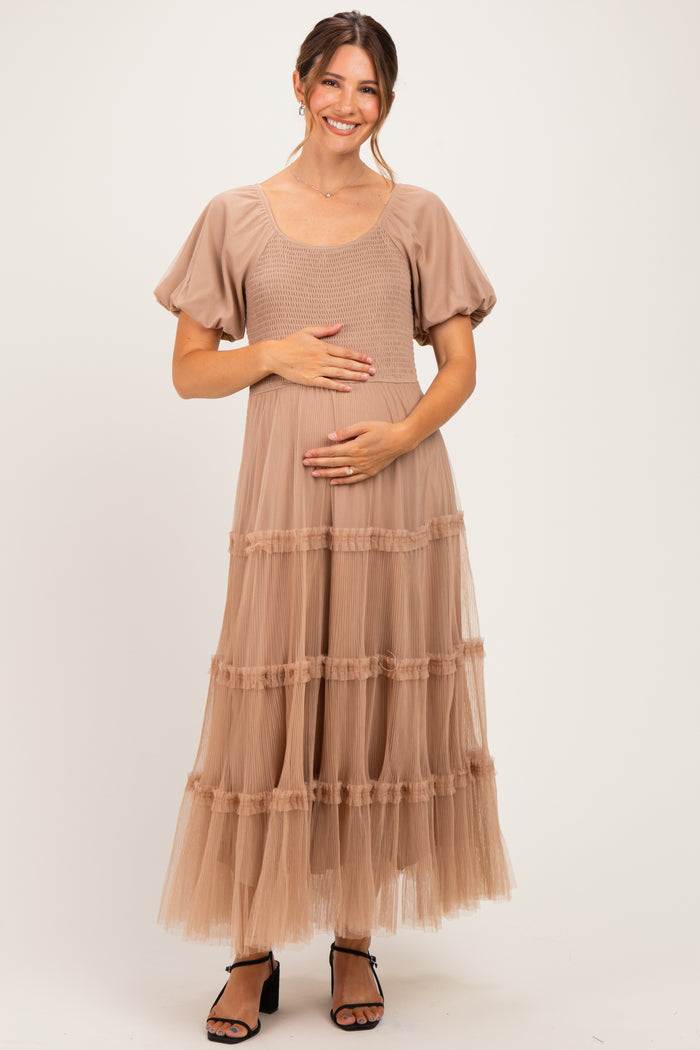 woman wearing a taupe maternity dress with puff sleeves and tiered tulle skirt, holding her baby bump
