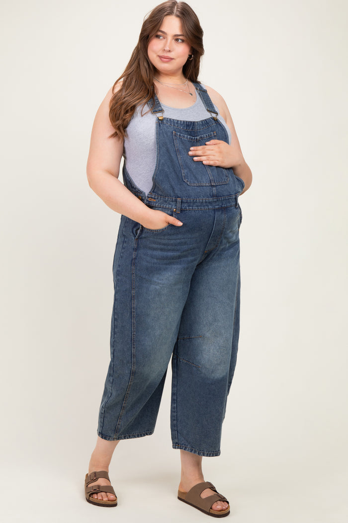 woman wearing a gray tank top and blue maternity plus denim overalls, holding her baby bump