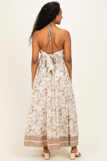 woman wearing a backless cream and beige floral maternity maxi dress with a tied halter neck and flowing silhouette