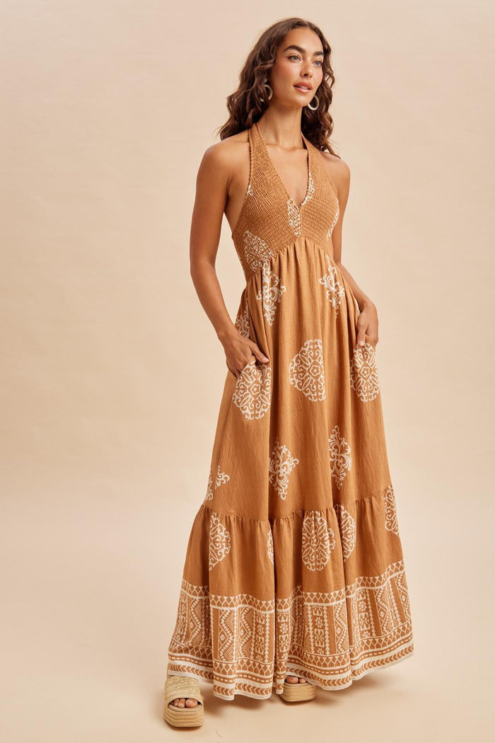 woman wearing a brown maxi dress with white intricate patterns and pockets, standing against a neutral background