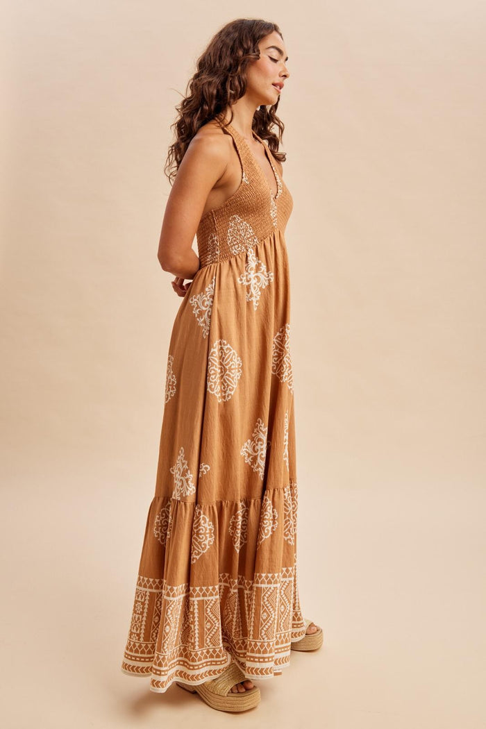 woman wearing a tan maxi dress with white floral patterns and a tiered skirt, standing sideways against a neutral background