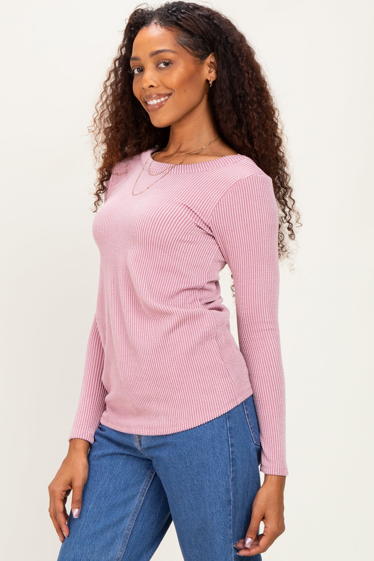 woman wearing a light pink ribbed long sleeve top paired with blue jeans, smiling and posing sideways